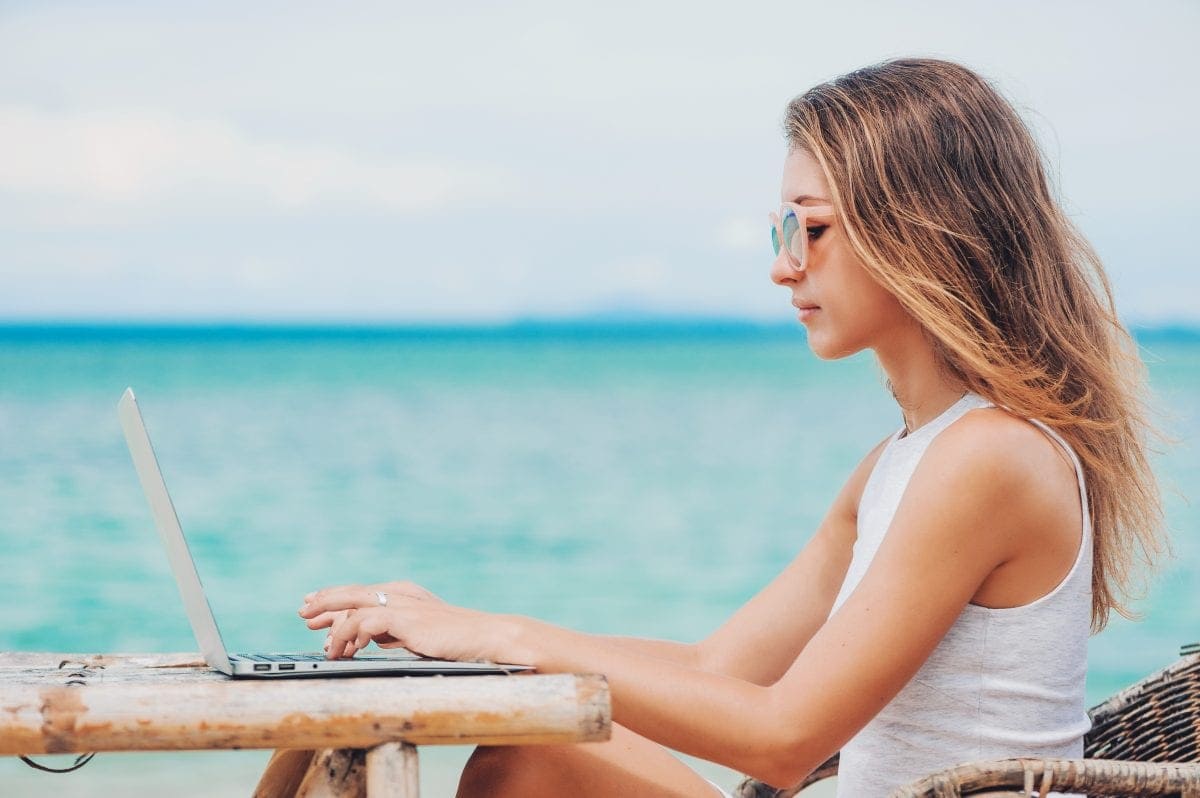 Young woman freelancer in dress sitting at the table on ocean background, using laptop on the beach.