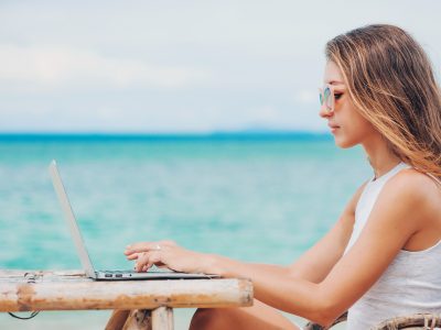 Young woman freelancer in dress sitting at the table on ocean background, using laptop on the beach.